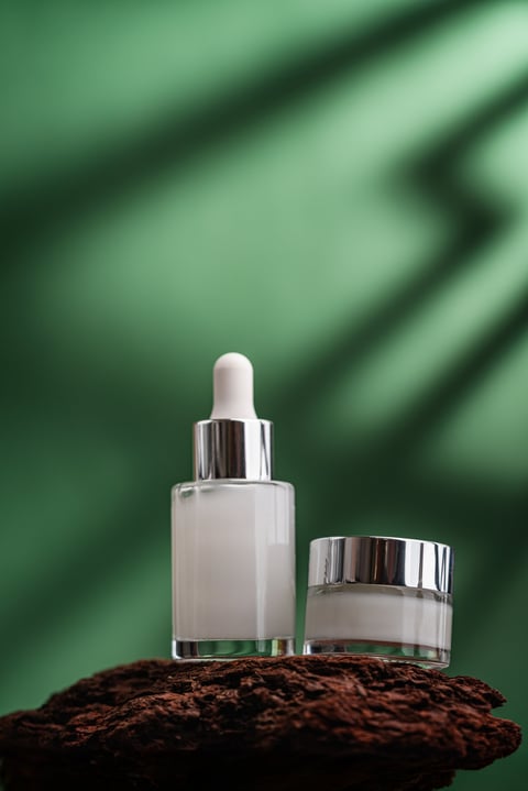 Two elegant glass cosmetic containers with skincare products placed on natural wooden texture against a green background. Minimalistic composition emphasizing beauty, purity, and wellness.
